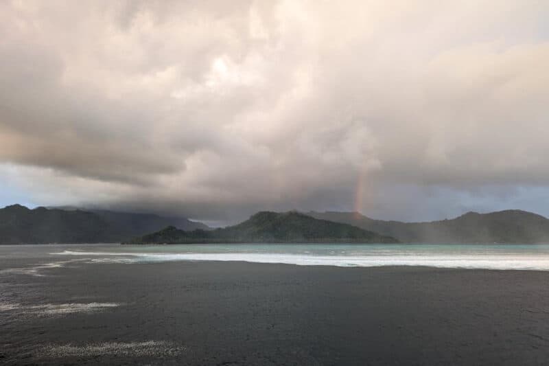 Weiße Wellenkämme, dichte Regenwolken und ein Regenbogen über dem Saumriff von Tahaa