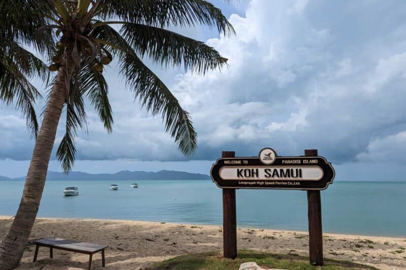 Ein Holzschild mit der Aufschrift Koh Samui am Strand mit einer Palme und Booten im Hintergrund