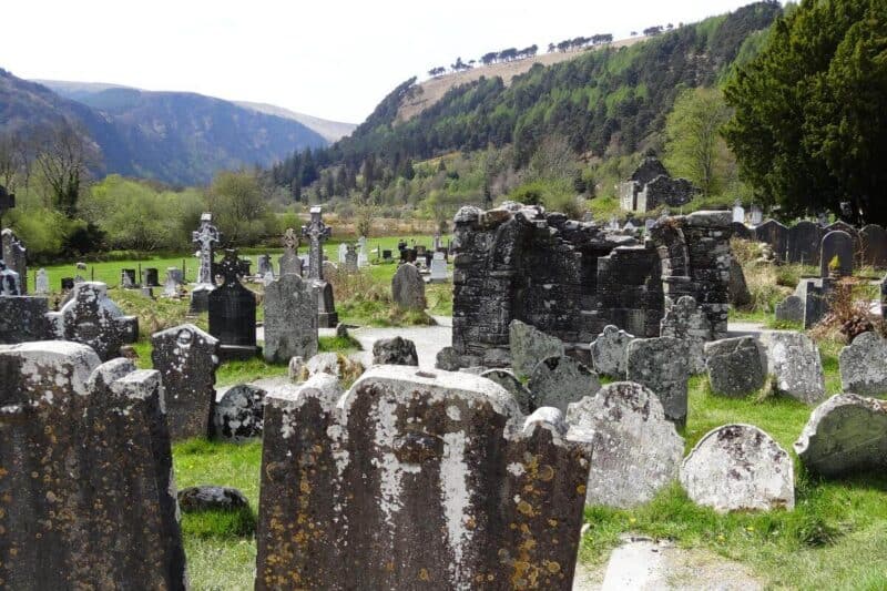 Grabkreuze und eine Kapellenruine vor den grünen Hügeln des Glendalough-Tals
