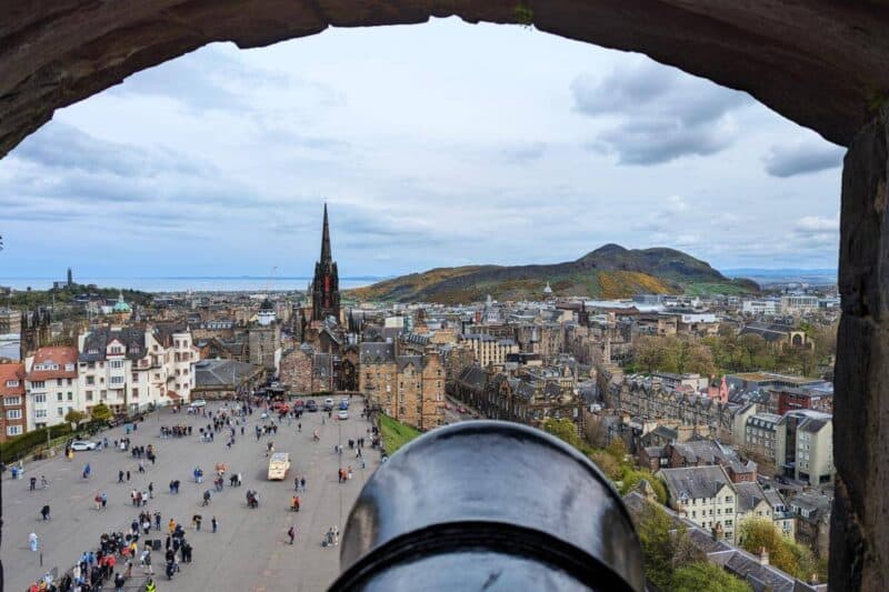 Aussicht durch eine Schießscharte des Edinburgh Castle mit Kanone im Vordergrund und der Altstadt von Edinburgh sowie dem Arthur's Seat im Hintergrund