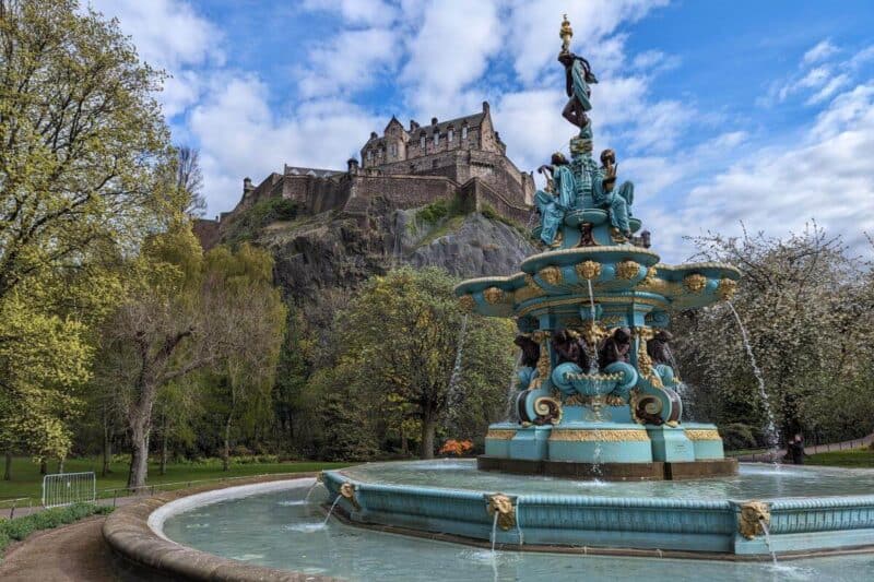 Ein blau und gold verzierter Springbrunnen vor dem Castle Rock mit dem Edinburgh Castle in den Princes Street Gardens